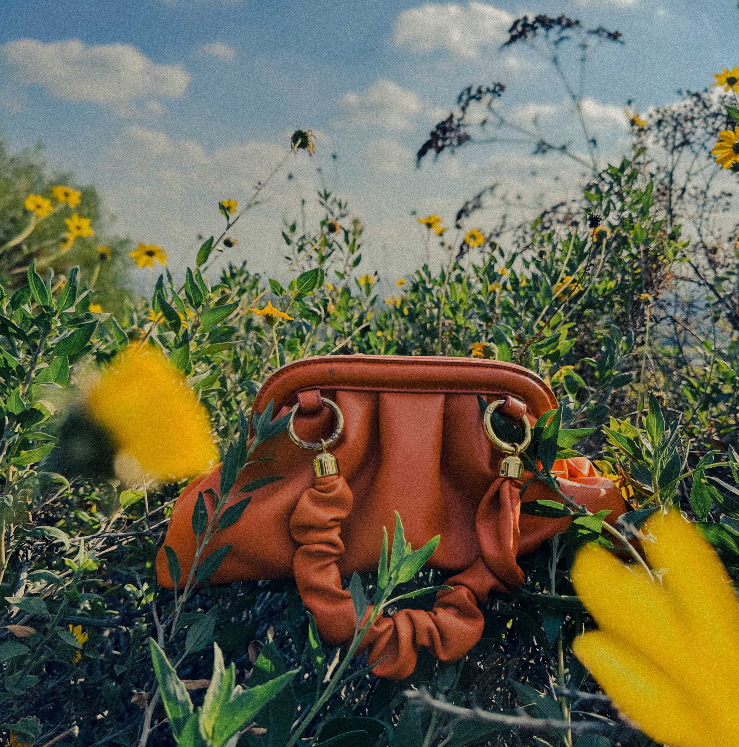 An orange vegan leather Reformation bag sits amongst yellow flowers with a view of the sky filled with clouds
